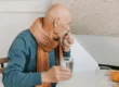 Elderly man sitting at a table with a glass of water, wearing a scarf, looking thoughtful. The image reflects the importance of hydration for the elderly and the impact of dehydration on cognitive health.