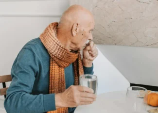 Elderly man sitting at a table with a glass of water, wearing a scarf, looking thoughtful. The image reflects the importance of hydration for the elderly and the impact of dehydration on cognitive health.