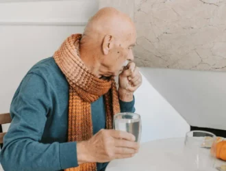 Elderly man sitting at a table with a glass of water, wearing a scarf, looking thoughtful. The image reflects the importance of hydration for the elderly and the impact of dehydration on cognitive health.