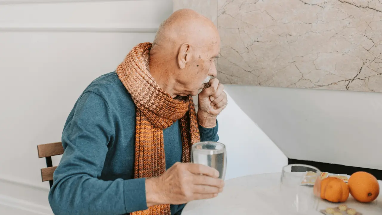 Elderly man sitting at a table with a glass of water, wearing a scarf, looking thoughtful. The image reflects the importance of hydration for the elderly and the impact of dehydration on cognitive health.