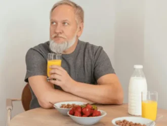 An elderly man with a beard sitting at a table, enjoying a healthy breakfast with a glass of orange juice, a bowl of strawberries, and a bowl of cereal with milk.