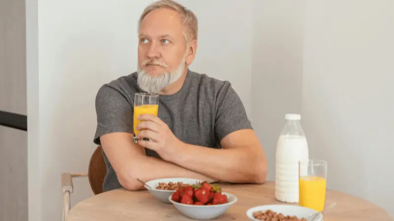 An elderly man with a beard sitting at a table, enjoying a healthy breakfast with a glass of orange juice, a bowl of strawberries, and a bowl of cereal with milk.