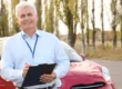 Senior driving instructor with clipboard, standing by a red car, illustrating new drivers license rules for seniors