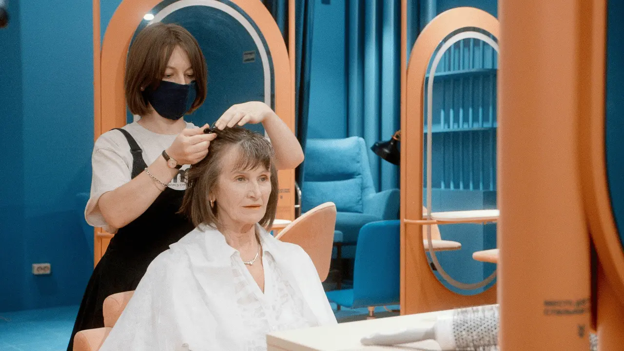 A senior woman receiving a stylish haircut in a modern salon, showcasing a low-maintenance hairstyle suitable for thin hair. A hairdresser is trimming her hair, helping her achieve a fuller, youthful look with minimal effort
