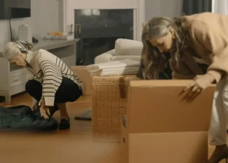 Two senior women, one with gray hair and the other with longer hair, are decluttering and organizing their home. One woman is bending down to pack a suitcase, while the other is sorting through items in a cardboard box. The scene captures the emotional and practical aspects of decluttering and hoarding help for seniors.