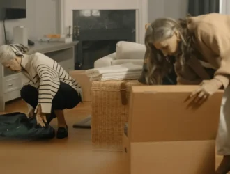 Two senior women, one with gray hair and the other with longer hair, are decluttering and organizing their home. One woman is bending down to pack a suitcase, while the other is sorting through items in a cardboard box. The scene captures the emotional and practical aspects of decluttering and hoarding help for seniors.