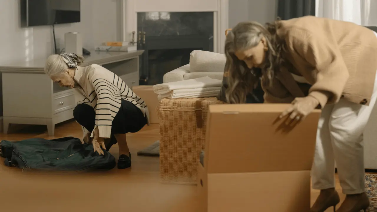 Two senior women, one with gray hair and the other with longer hair, are decluttering and organizing their home. One woman is bending down to pack a suitcase, while the other is sorting through items in a cardboard box. The scene captures the emotional and practical aspects of decluttering and hoarding help for seniors.