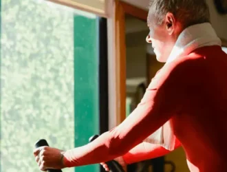 An elderly man using an elliptical machine, wearing a red shirt and a towel around his neck, while enjoying a workout session. The natural light from a nearby window highlights his focused expression as he engages in the exercise.
