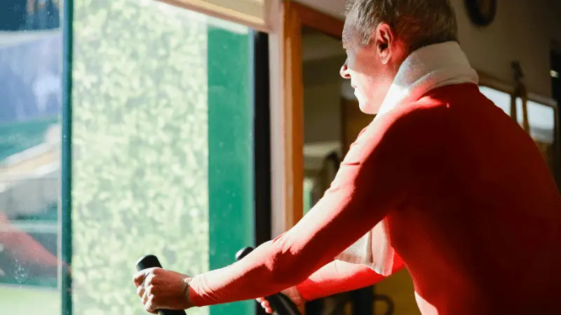 An elderly man using an elliptical machine, wearing a red shirt and a towel around his neck, while enjoying a workout session. The natural light from a nearby window highlights his focused expression as he engages in the exercise.